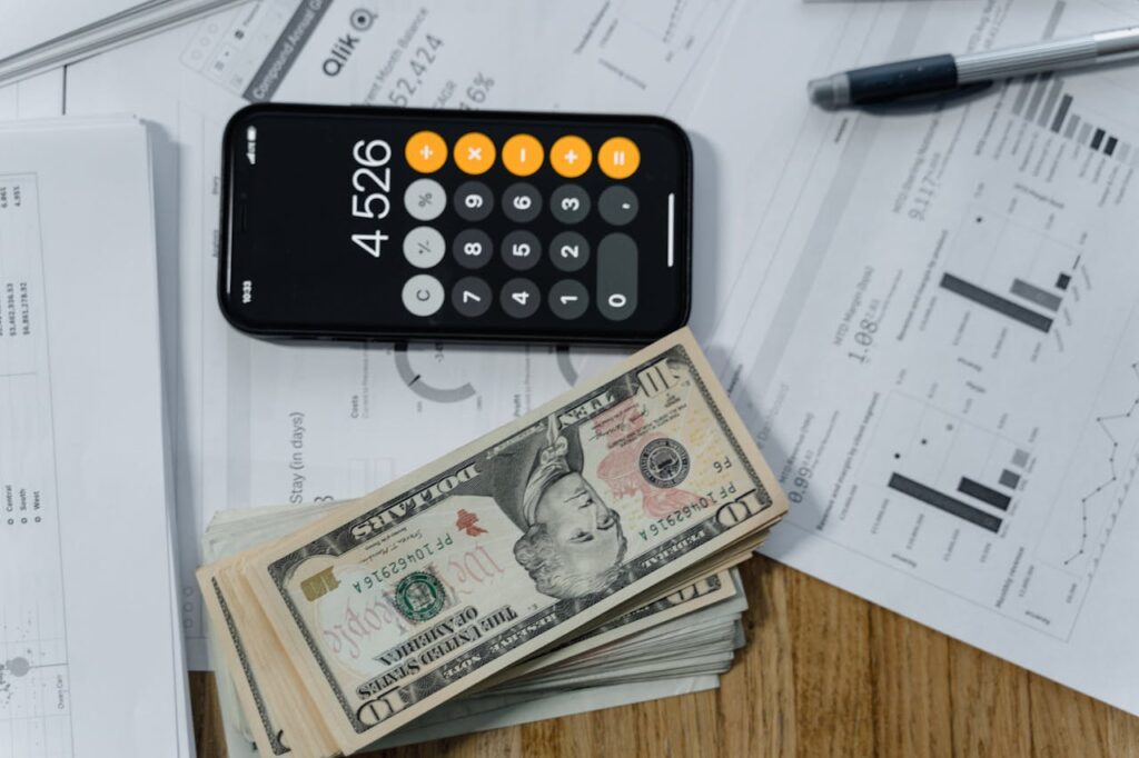 A top-down view of a desk with investment statements, an iPhone calculator, and a stack of USD cash for a homebuyer considering asset based finance. 