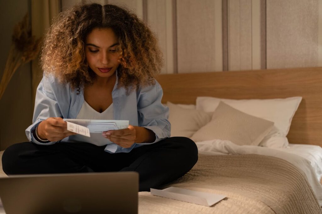 A woman is sitting on her bed reading her mortgage statement she received in the mail in front of her laptop. 