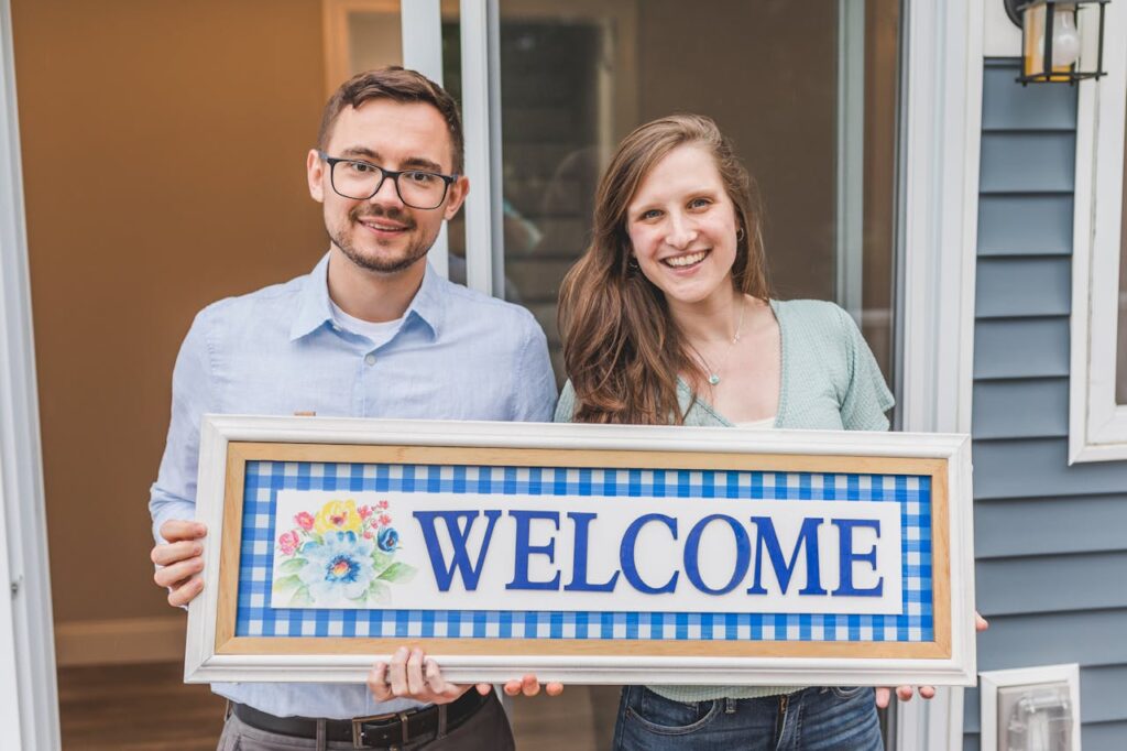 A couple is holding a "welcome" sign in front of their new home after figuring out where to get a mortgage.